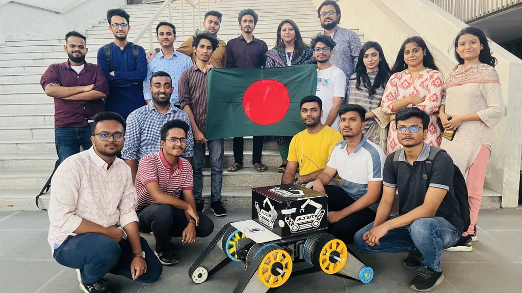 The BRACU Alter squad — about seventeen people — arranged on a broad flight of steps, holding a Bangladesh flag between them. On the ground in front sits the rescue rover: a black box body lettered ALTER, carried on four tracked wheels with yellow and blue sprockets, with two further track arms extending forward from the chassis.